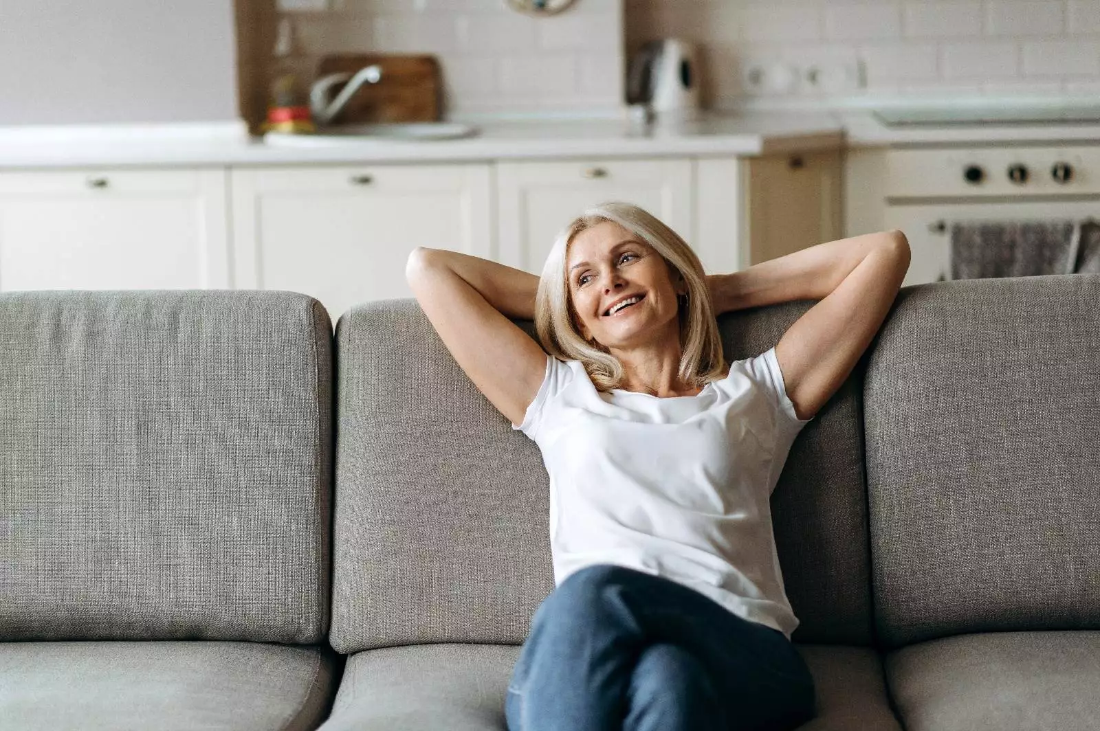 A woman with blonde hair, wearing a white shirt and jeans, sits on a gray couch in a bright kitchen, smiling and relaxing with her hands behind her head, embodying the sense of calm that many seek through SPRAVATO™ Treatment.