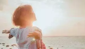 Person with short hair stands by the ocean at sunset, arms outstretched, basking in the sunlight with a peaceful expression. The sun shines brightly behind them.