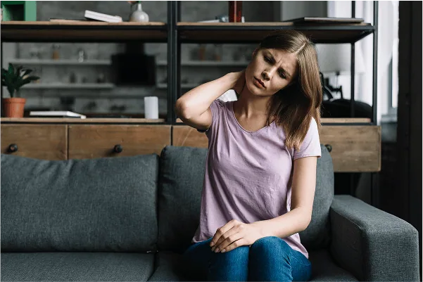 chronic-pain-01 A woman sitting on a gray couch holds her neck with one hand, appearing to be in pain or discomfort—possibly experiencing chronic pain. She is wearing a light purple shirt and blue jeans in a modern living room setting.