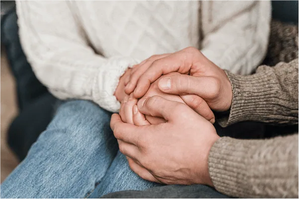 chronic-pain-02 Two people sit closely together, one wearing a white sweater and the other a brown sweater, gently holding hands in a comforting gesture—a quiet moment of support for someone living with chronic pain.
