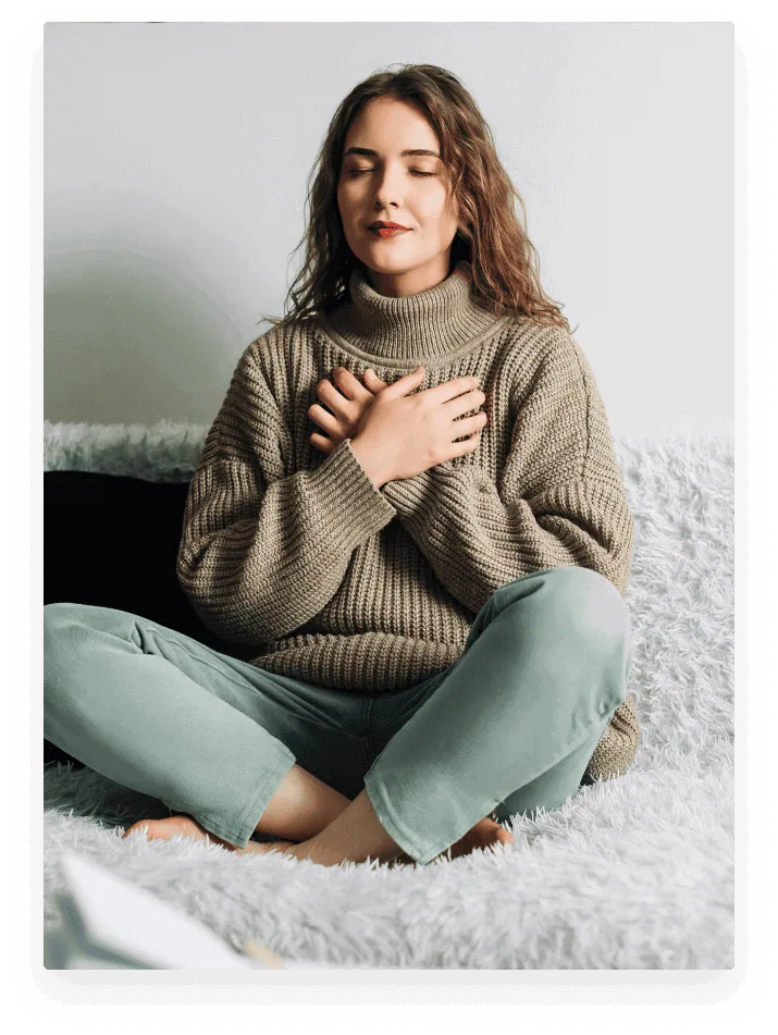 girl-sitting-in-yoga-pose A woman with long hair sits cross-legged on a couch, eyes closed, hands crossed over her chest, wearing a chunky knit sweater and light pants, appearing calm and relaxed.